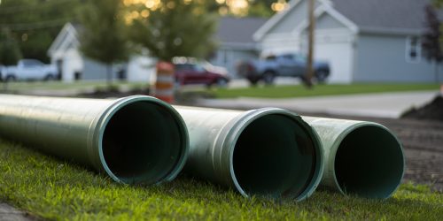 Large blue pipe sits on the grass next to a road and drain repair project in a residential neighborhood.